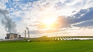 Foto, Blick auf eine grüne Wiese mit einer großen Photovolataik-Anlage, im Hintergrund ein Kohlekraftwerk sowie Windräder.