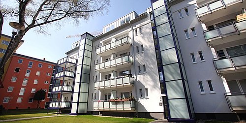 Foto, Mehrstöckiges Wohngebäude mit Balkonen und Aufzügen, umgeben von Rasen und Bäumen bei klarem Himmel