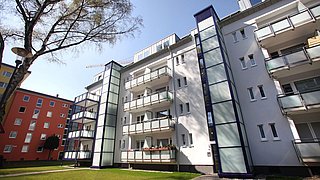 Foto, Mehrstöckiges Wohngebäude mit Balkonen und Aufzügen, umgeben von Rasen und Bäumen bei klarem Himmel