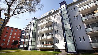 Foto, Mehrstöckiges Wohngebäude mit Balkonen und Aufzügen, umgeben von Rasen und Bäumen bei klarem Himmel