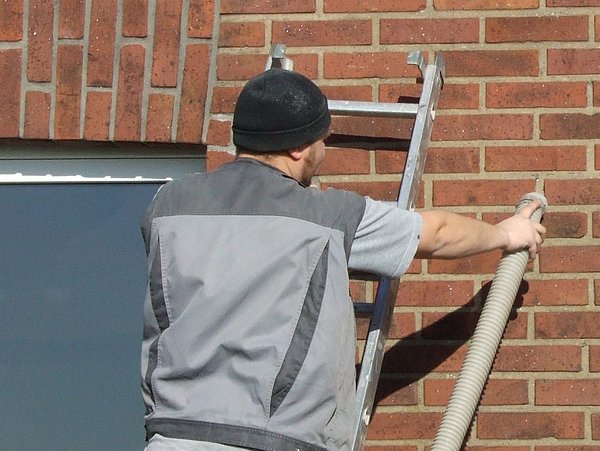Foto, Handwerker mit schwarzer Mütze und grauer Arbeitskleidung hält eine Leiter und einen Schlauch an eine rote Backsteinwand.