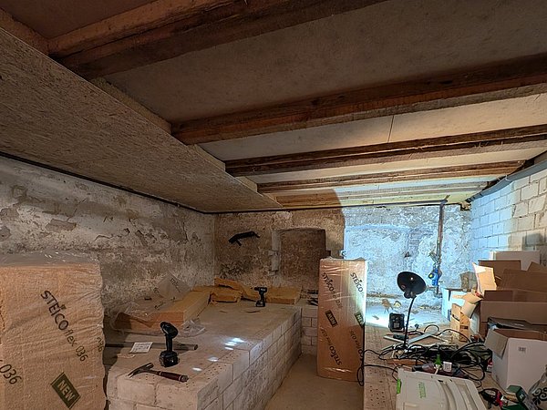 Foto, Keller mit sichtbarer Decke aus Holzbalken, Dämmplatten und verschiedenen Werkzeugen sowie Kartons auf dem Boden