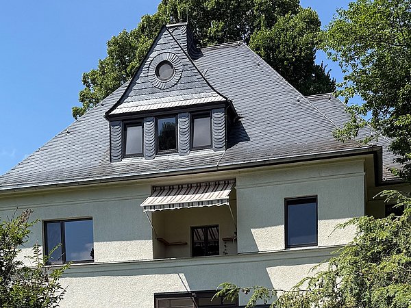 Foto, Im Vordergrund steht ein freistehendes Wohnhaus mit zwei Stockwerken und einem ausgebauten Dachgeschoss. Die Fassade ist hell und das Dach mit dunklen Schieferplatten gedeckt. Über dem Eingangsbereich befindet sich ein kleiner Wintergarten mit Glasdach und gemauertem Sockel aus rötlichem Naturstein.