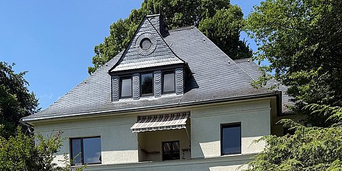 Foto, Im Vordergrund steht ein freistehendes Wohnhaus mit zwei Stockwerken und einem ausgebauten Dachgeschoss. Die Fassade ist hell und das Dach mit dunklen Schieferplatten gedeckt. Über dem Eingangsbereich befindet sich ein kleiner Wintergarten mit Glasdach und gemauertem Sockel aus rötlichem Naturstein.