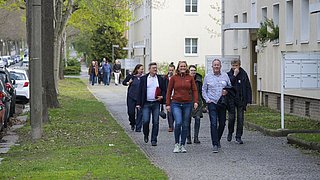 Foto, Gruppe von Menschen geht auf einem gepflasterten Gehweg neben einem Wohngebäude und einer Baumreihe