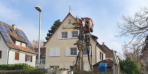 Foto, im Vordergrund befindet sich ein Lastwagen mit Baumaterialien, dahinter ein Spezialgerät zur Erdwärmebohung, im Hintergrund ein Einfamilienhaus.