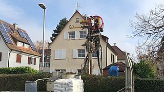 Foto, im Vordergrund befindet sich ein Lastwagen mit Baumaterialien, dahinter ein Spezialgerät zur Erdwärmebohung, im Hintergrund ein Einfamilienhaus.