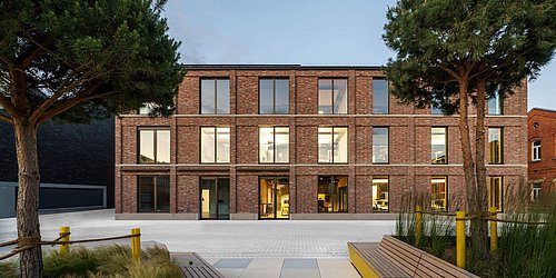 Foto, Ein modernes, dreistöckiges Gebäude mit einer Fassade aus rotbraunem Klinker steht im Mittelpunkt. Große Fensterflächen prägen die gleichmäßige Struktur. Im Vordergrund befinden sich helle Sitzbänke aus Holz, Pflanzflächen mit Gräsern und zwei Bäume, die den Eingangsbereich rahmen.