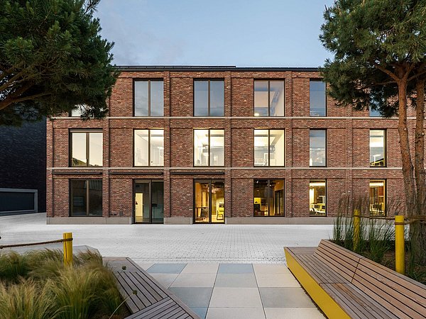 Foto, Ein modernes, dreistöckiges Gebäude mit einer Fassade aus rotbraunem Klinker steht im Mittelpunkt. Große Fensterflächen prägen die gleichmäßige Struktur. Im Vordergrund befinden sich helle Sitzbänke aus Holz, Pflanzflächen mit Gräsern und zwei Bäume, die den Eingangsbereich rahmen.