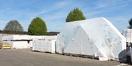 Foto, mit Folie umwickelte Gebäude-Module im Freien unter klarem Himmel mit Bäumen im Hintergrund.