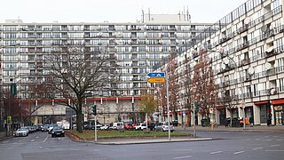 Foto, Ein großes Wohngebäude mit vielen Balkonen erstreckt sich über die gesamte Bildbreite. Im Vordergrund befindet sich eine Straßenkreuzung mit mehreren Fahrspuren, parkenden Autos und einem begrünten Mittelstreifen. Zwei Straßenschilder mit blauer und gelber Beschriftung stehen auf einer Verkehrsinsel. Links und rechts säumen weitere Wohnhäuser die Straße. Einige Bäume ohne Laub stehen am Straßenrand. Die Szene wirkt städtisch und dicht bebaut.