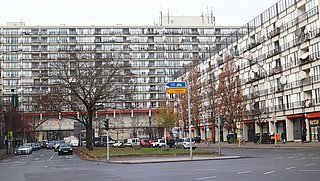 Foto, Ein großes Wohngebäude mit vielen Balkonen erstreckt sich über die gesamte Bildbreite. Im Vordergrund befindet sich eine Straßenkreuzung mit mehreren Fahrspuren, parkenden Autos und einem begrünten Mittelstreifen. Zwei Straßenschilder mit blauer und gelber Beschriftung stehen auf einer Verkehrsinsel. Links und rechts säumen weitere Wohnhäuser die Straße. Einige Bäume ohne Laub stehen am Straßenrand. Die Szene wirkt städtisch und dicht bebaut.
