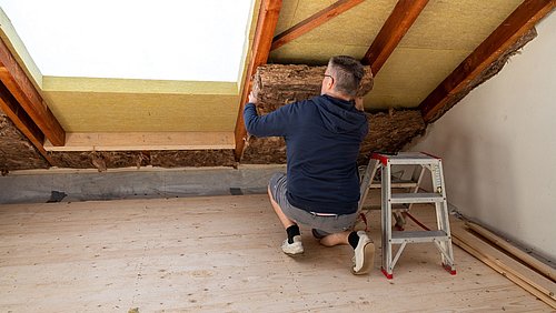Foto, Person kniet auf dem Boden eines Dachbodens und bringt Dämmmaterial zwischen die Dachbalken ein.