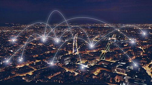 Foto, Luftaufnahme einer nächtlichen Stadt mit beleuchteten Netzleitungen, die sich über die Gebäude wölben und auf die digitale Konnektivität hinweisen.
