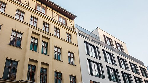 Foto, Blick auf zwei Gebäude, ein sanierter Altbau und ein Neubau mit modernem Glasdesign, unter einem klaren blauen Himmel.