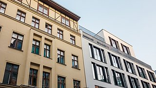 Foto, Blick auf zwei Gebäude, ein sanierter Altbau und ein Neubau mit modernem Glasdesign, unter einem klaren blauen Himmel.