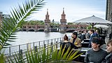 Foto, Menschen sitzen auf einer Terrasse am Wasser mit Blick auf die Oberbaumbrücke in Berlin bei bewölktem Himmel