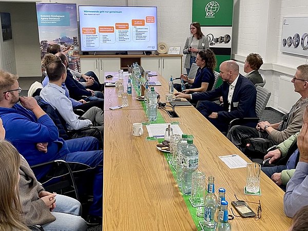 Foto, Menschen sitzen an langem Tisch, der zu einem Raumende ausgerichtet ist. Vorne werden Folien über die Wärmewende gezeigt und präsentiert.