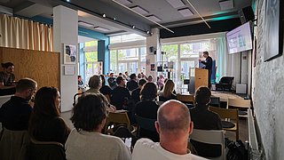 Foto, Blick in einen hellen Seminarraum mit mehreren sitzenden Personen, die einem Vortragenden an einem Rednerpult zuhören.
