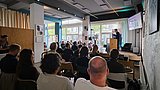 Foto, Blick in einen hellen Seminarraum mit mehreren sitzenden Personen, die einem Vortragenden an einem Rednerpult zuhören.