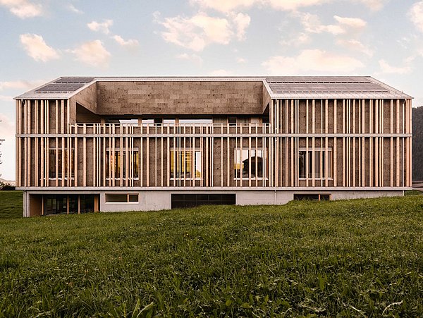 Foto, modernes, langgestrecktes Gebäude mit Holzfassade und großen Fenstern auf einer grünen Wiese unter bewölktem Himmel