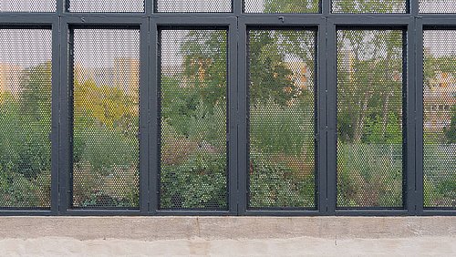 Foto, Fensterfront mit schwarzem Metallrahmen und perforierten Metallgittern vor einer grünen Baumkulisse.