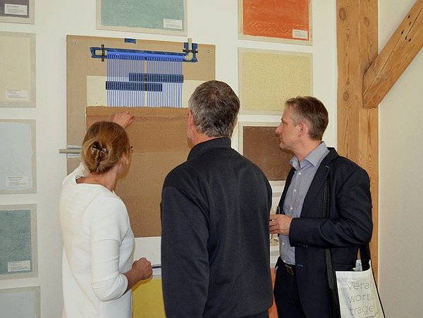 Foto, Drei Personen betrachten und diskutieren Muster von Wandbelägen in einem Ausstellungsraum der Naturbauschule