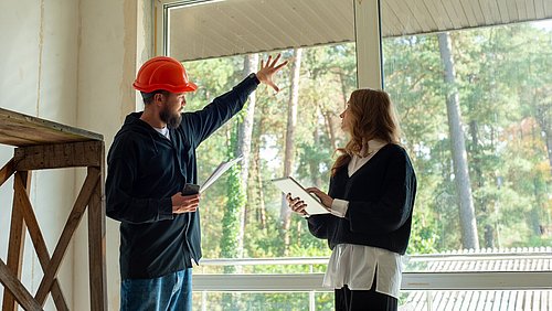 Foto, ein Mann mit orangefarbener Sicherheitshelm zeigt mit der Hand auf ein Fenster, während eine Frau mit einem Tablet in der Hand ihm zuhört. Im Hintergrund sind Bäume sichtbar.