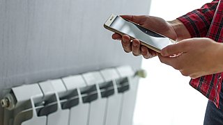 Foto, Hand hält Smartphone vor Heizkörper an weißer Wand.