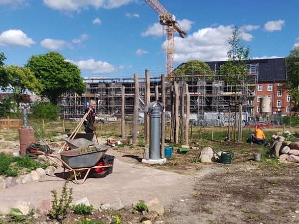 Foto, Im Vordergrund befindet sich eine Baustelle mit losem Erdreich, einer Schubkarre und verschiedenen Werkzeugen. Zwei Personen arbeiten im Gelände, eine steht mit einer Schaufel, die andere sitzt neben einem Steinhaufen. Im Hintergrund sind Baugerüste und ein Kran zu sehen, dahinter mehrstöckige Gebäude. Der Himmel ist blau mit wenigen weißen Wolken, Bäume spenden Schatten.