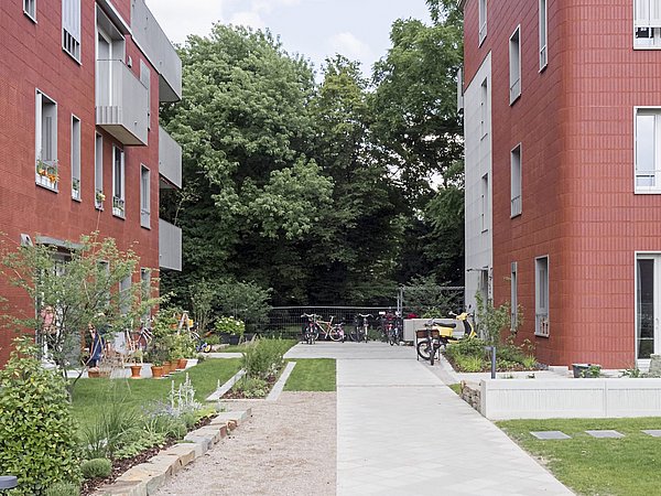 Zwei moderne Wohngebäude mit roter Fassade, umgeben von Gärten. In der Mitte ein gepflasterter Weg, links Fahrräder und ein Motorrad sichtbar.