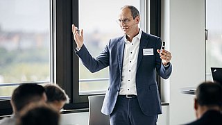 Foto, Mann in blauem Anzug und weißem Hemd hält Präsentation vor Publikum in hellem Raum mit großen Fenstern