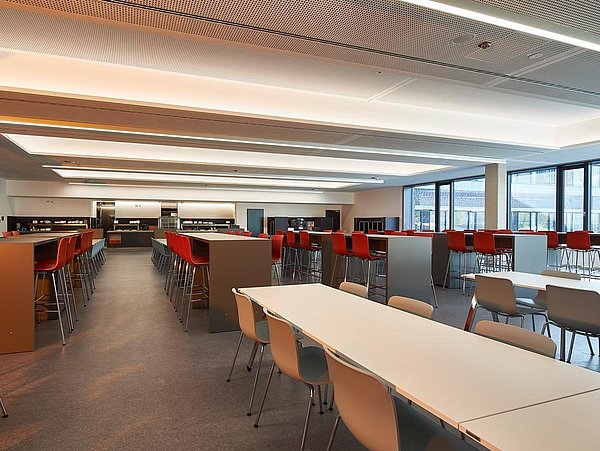 Foto, geräumige Caféteria mit vielen Tischen und Stühlen, die in Reihen stehen. Im hinteren Teil des Raumen sind Tische und Stühle höher als im vorderen Teil des Raumes.