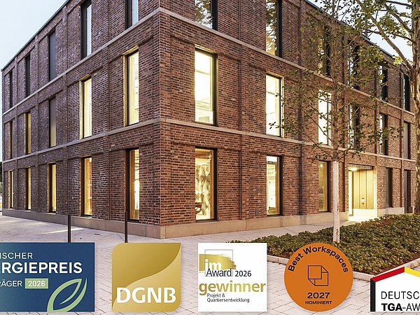 Foto, Ein modernes, dreistöckiges Gebäude aus dunkelrotem Backstein steht an einer gepflasterten Fläche. Die großen, rechteckigen Fenster sind gleichmäßig angeordnet und leuchten von innen. Links im Bild sind einige Straßenlaternen zu sehen, rechts wachsen mehrere hohe Bäume mit grünen Blättern, unten im Bild sind Siegel der Preise: Bayerischer Energiepreis 2026, DGNB, im Award gewinner 2026 Projekt und Quartiersentwicklung, Best Workspaces 2027, Deutscher TGA-Award. 