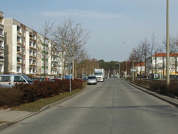 Foto, Blick auf eine breite Straße mit parkenden Fahrzeugen und mehrstöckigen Wohngebäuden auf der linken Seite sowie Bäumen ohne Blätter