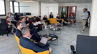 Foto, Mehrere Personen sitzen in einem modernen Büro auf gelben Sesseln und schwarzen Stühlen, während eine Person vorne stehend eine Präsentation hält.