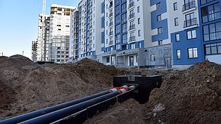 Foto, Baugrube mit mehreren großen schwarzen Rohren vor mehrstöckigen Wohngebäuden mit blauen Fassaden und einem Baukran im Hintergrund.