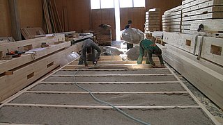 Foto, Zwei Personen verlegen Schafwolle als Dämmmaterial zwischen Holzbalken in einer Werkstatt mit Holzrahmenbau