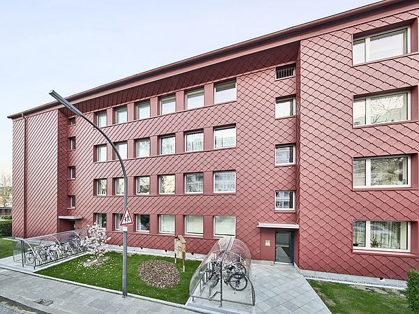 Foto, Mehrstöckiges Wohngebäude mit roten Fassadenplatten und mehreren Fenstern, Fahrradständer mit überdachtem Unterstand davor