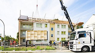 Foto, Blick von der Straße auf ein mehrstöckiges Wohngebäude. Das Dach liegt offen, ein Kran hebt ein vorgefertigtes Dachmodul in die Höhe.