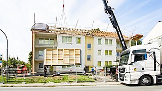 Foto, Blick von der Straße auf ein mehrstöckiges Wohngebäude. Das Dach liegt offen, ein Kran hebt ein vorgefertigtes Dachmodul in die Höhe.