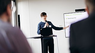 Foto, Mann steht mit Mikrofon vor Laptop und zeigt auf Präsentationsfolie mit Text zu Klimaneutralität.