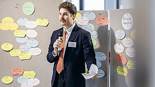 Foto, Mann im dunklen Anzug mit orangefarbener Krawatte hält Mikrofon und Papier vor Pinnwand mit bunten Moderationskarten.
