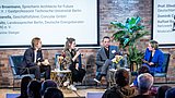 Foto, Vier Personen sitzen auf einer Bühne vor Publikum, diskutieren bei der Jahrestagung 2024, im Hintergrund Text mit Namen und Titeln der Panelteilnehmer