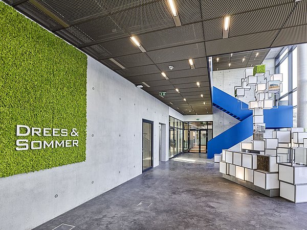 Foto, hoher Innenraum mit teilweise begrünter Wand links und einer geometrischen Skulpture auf der rechten Seite