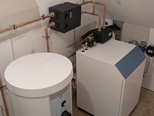 Foto, Heizungsanlage mit rundem Warmwasserspeicher und rechteckiger Wärmepumpe, verbunden durch Kupferrohre in einem Raum mit weiß gestrichenen Wänden und Holzboden