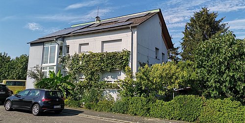Foto, Zweistöckiges Wohnhaus mit Solarpaneelen auf dem Dach, umgeben von grünen Pflanzen und parkenden Autos an der Straße