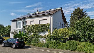 Foto, Zweistöckiges Wohnhaus mit Solarpaneelen auf dem Dach, umgeben von grünen Pflanzen und parkenden Autos an der Straße