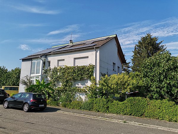 Foto, Zweistöckiges Wohnhaus mit Solarpaneelen auf dem Dach, umgeben von grünen Pflanzen und parkenden Autos an der Straße