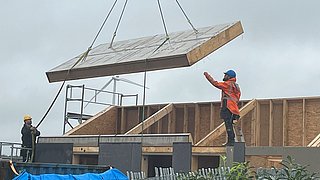 Foto, zwei Bauarbeiter mit Schutzhelmen und Warnkleidung heben mit einem Kran ein großes, rechteckiges Bauteil über einem Rohbau an.